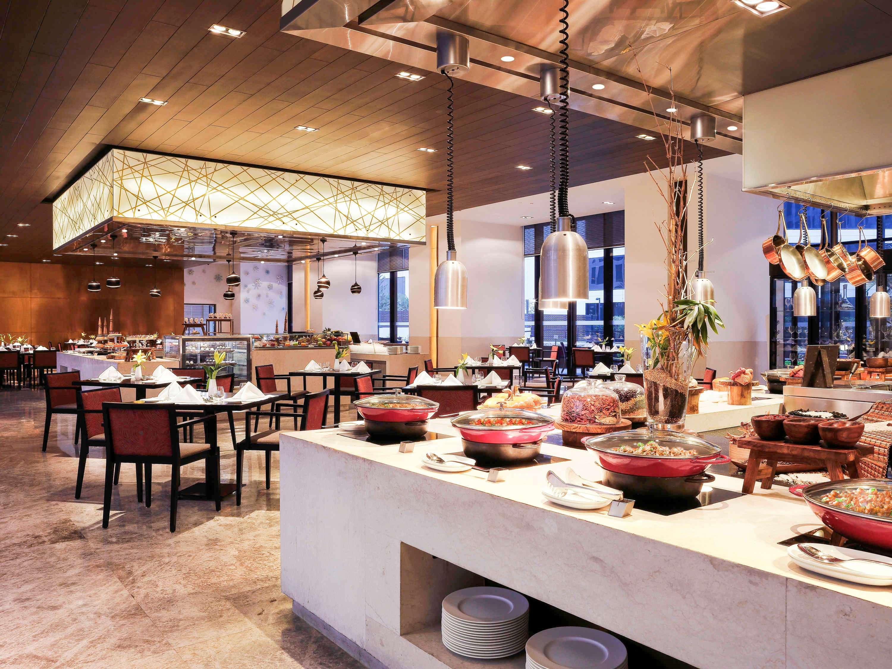 노보텔 뉴델리 인터내셔널 에어포트 언 아코르 호텔 브랜드 외부 사진 The photo shows a modern restaurant interior. There is a buffet area featuring various dishes, with several red bowls and an assortment of food items. The dining area includes tables and chairs with a sleek design, and the ambiance is bright and invi
