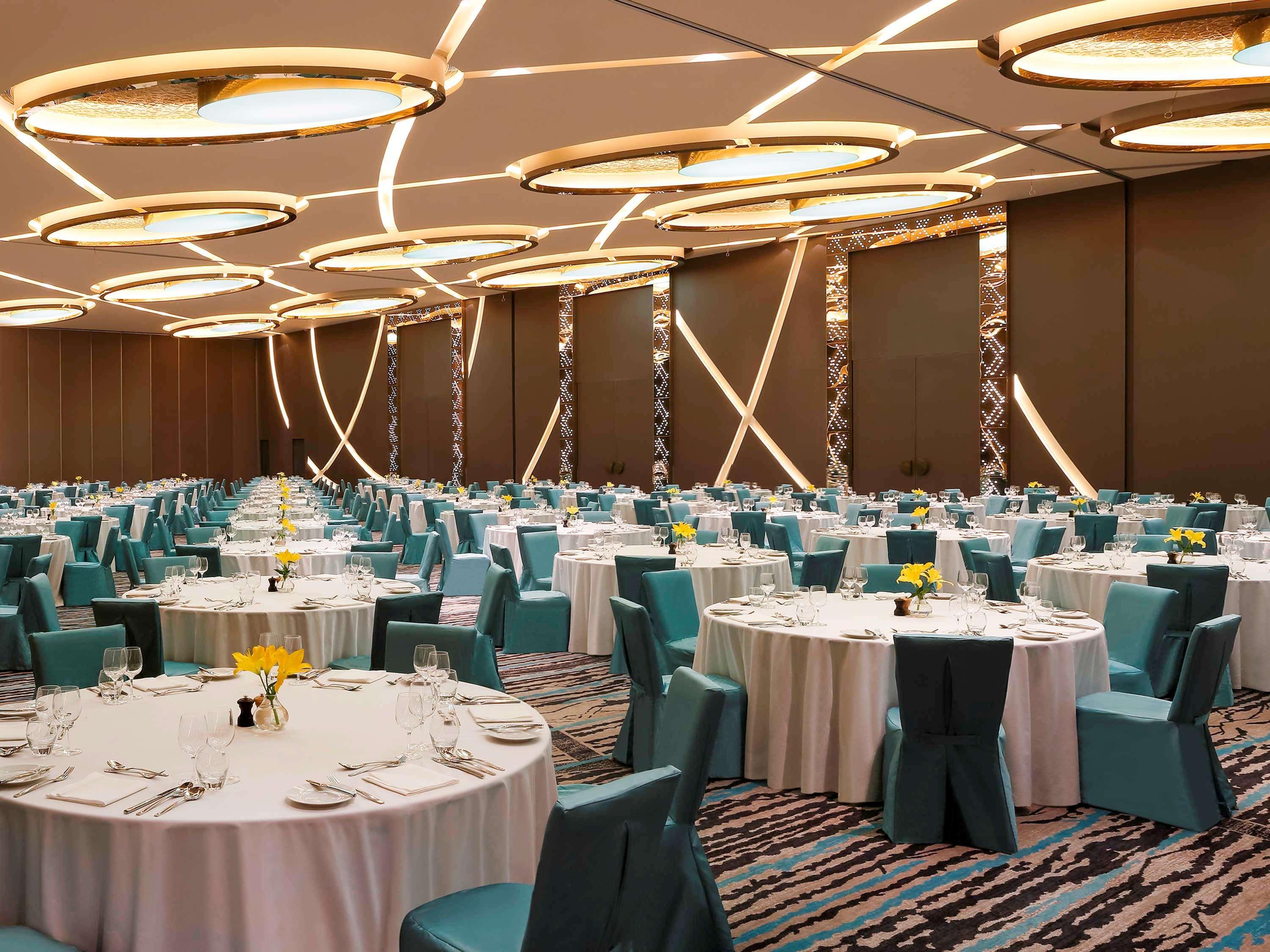 노보텔 뉴델리 인터내셔널 에어포트 언 아코르 호텔 브랜드 외부 사진 The photo depicts a spacious banquet hall set up for an event. There are multiple round tables covered with white tablecloths, each set with dining utensils, plates, and small vases containing yellow flowers. The chairs are upholstered in a teal fabr