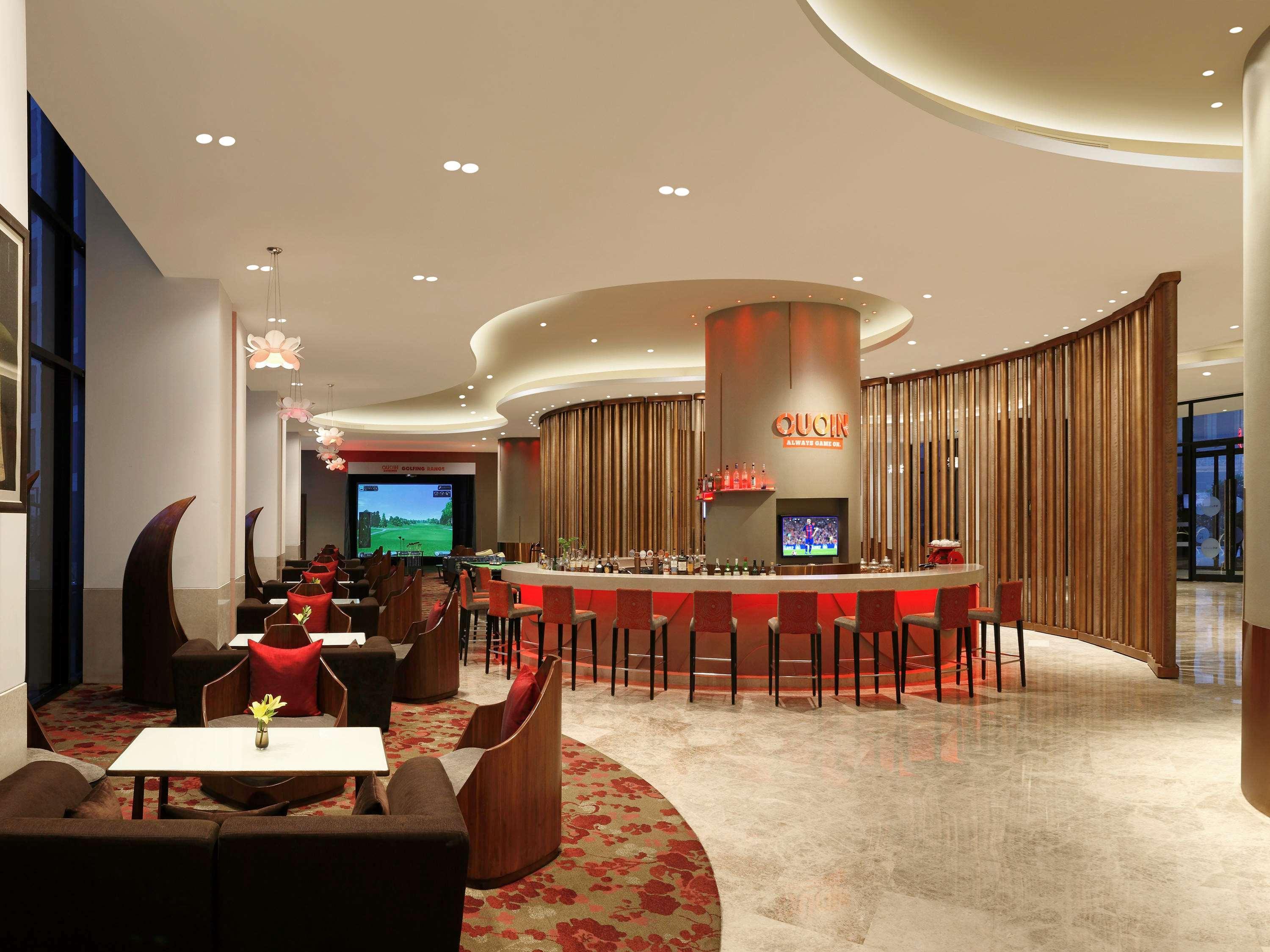 노보텔 뉴델리 인터내셔널 에어포트 언 아코르 호텔 브랜드 외부 사진 The photo shows a modern and stylish interior of a lounge or bar. The space features a curved bar in the center, surrounded by seating arrangements that include dark-colored couches and chairs. The design elements include a mix of smooth surfaces and