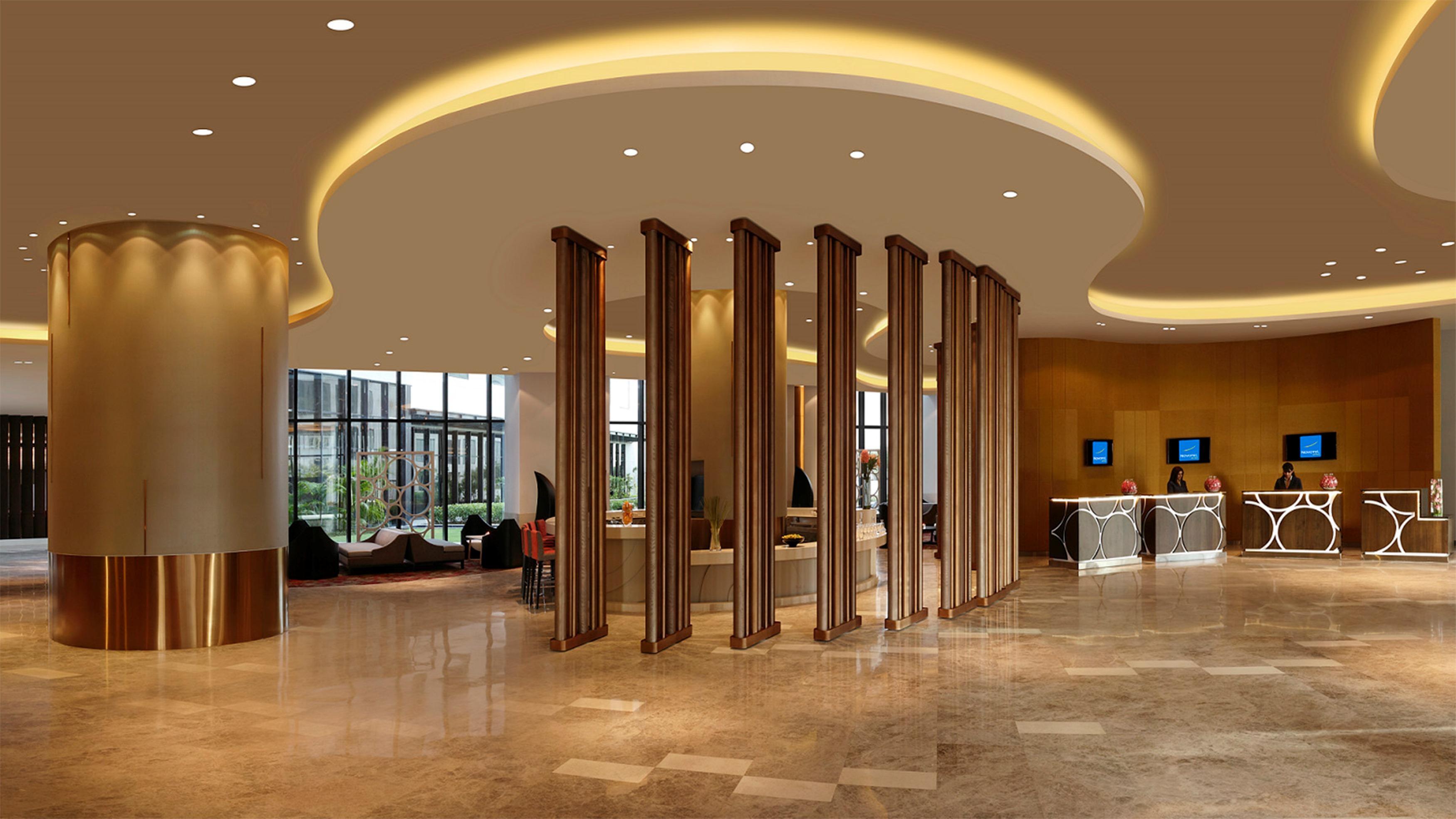 노보텔 뉴델리 인터내셔널 에어포트 언 아코르 호텔 브랜드 외부 사진 The photo shows a modern hotel lobby with a spacious and elegant design. The ceiling features a smooth, curved shape with ambient lighting creating a warm atmosphere. In the center, there are vertical wooden elements that add a sense of structure and