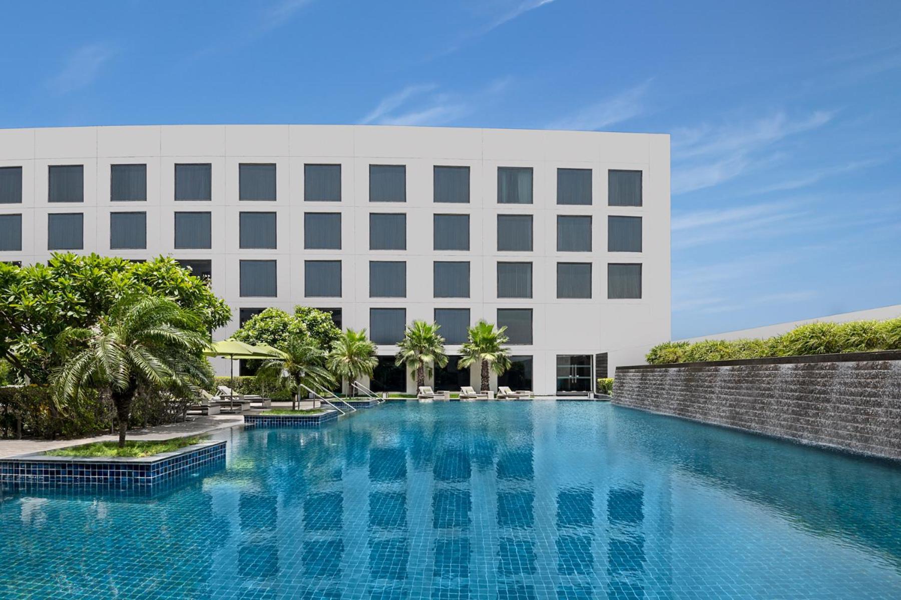 노보텔 뉴델리 인터내셔널 에어포트 언 아코르 호텔 브랜드 외부 사진 The photo shows a modern hotel building with a sleek, minimalist design. In front of the hotel is a large swimming pool, which reflects the blue sky and the building. Surrounding the pool are palm trees and lounging areas, creating a relaxing atmosph