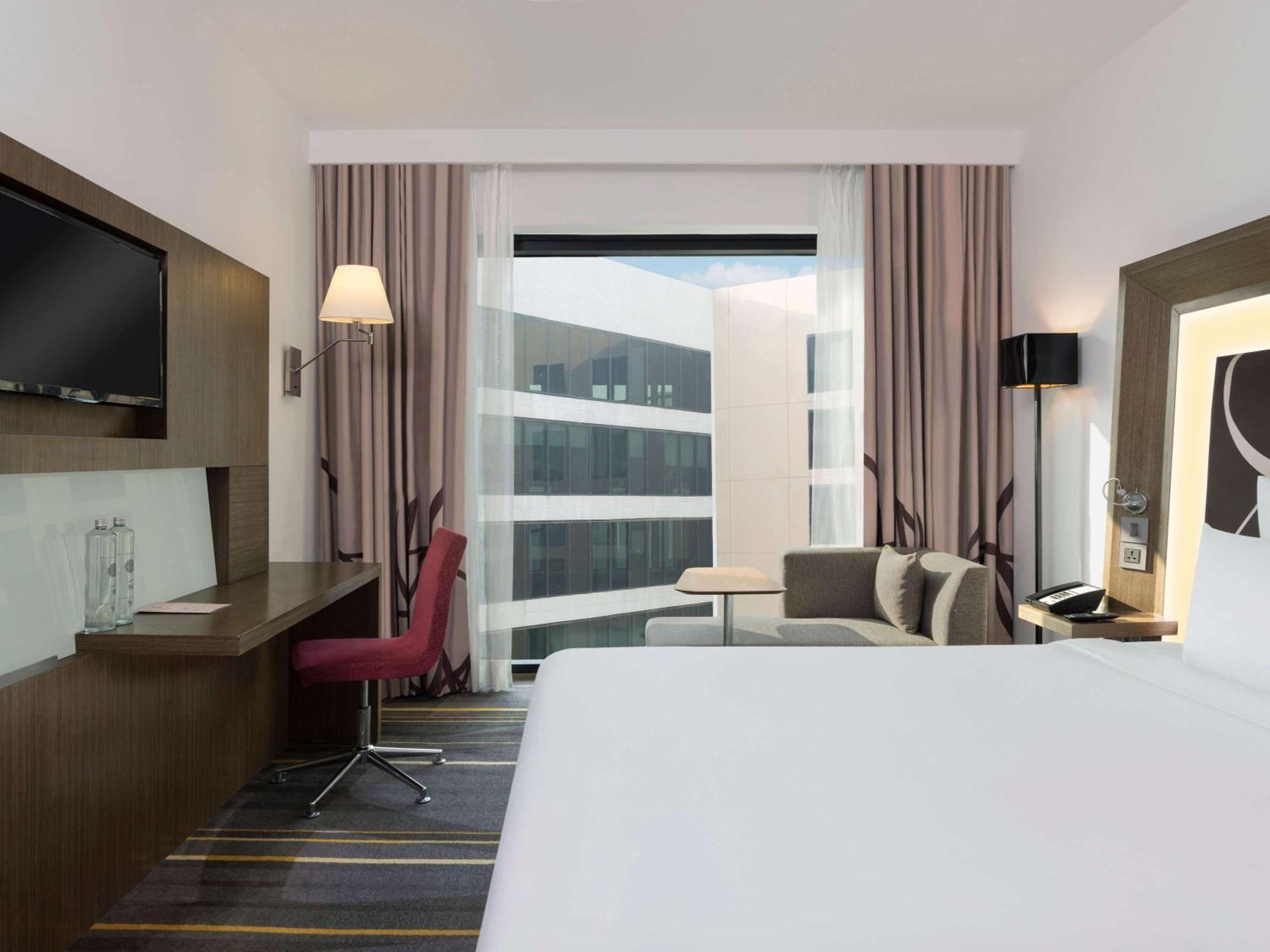 노보텔 뉴델리 인터내셔널 에어포트 언 아코르 호텔 브랜드 외부 사진 The photo shows a modern hotel room. It features a spacious layout with a large bed in the foreground. There is a window with sheer curtains that lets in natural light, revealing a view of a building outside. To one side, there's a small seating area