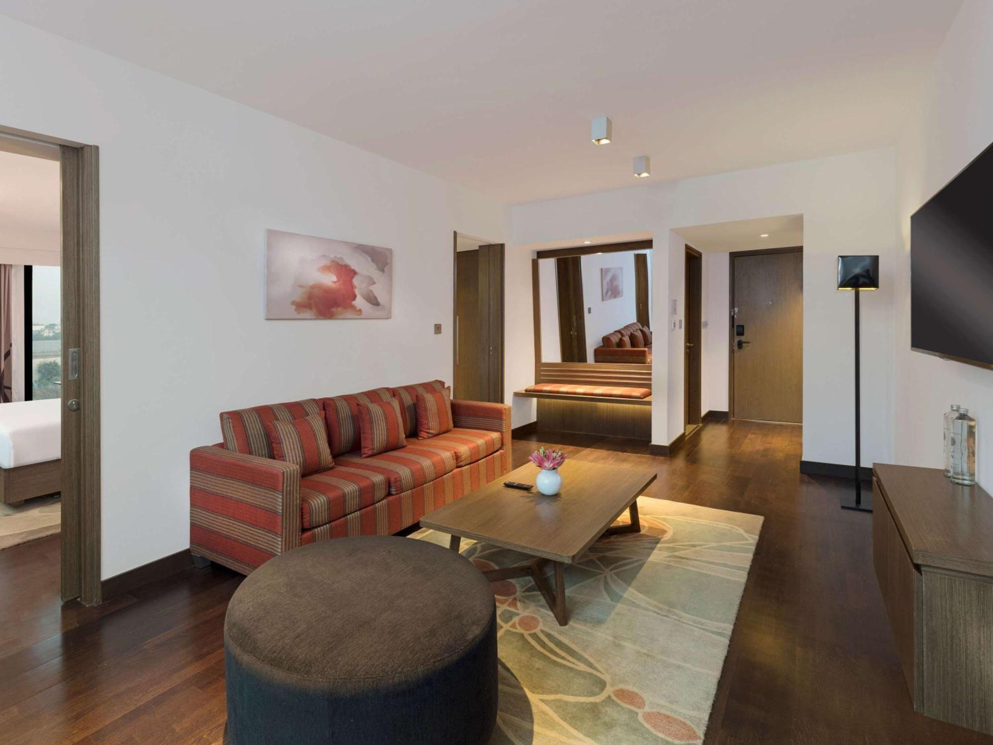 노보텔 뉴델리 인터내셔널 에어포트 언 아코르 호텔 브랜드 외부 사진 The photo shows a modern hotel room or suite. It features a spacious living area with a striped sofa and a round ottoman. There is a wooden coffee table in the center, and a decorative rug on the floor. The walls are painted in light colors, and ther